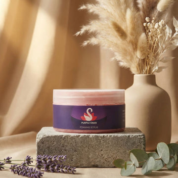 Jar of 'Purple Swan' foaming scrub on a stone block with a vase of dried flowers and lavender in the background.