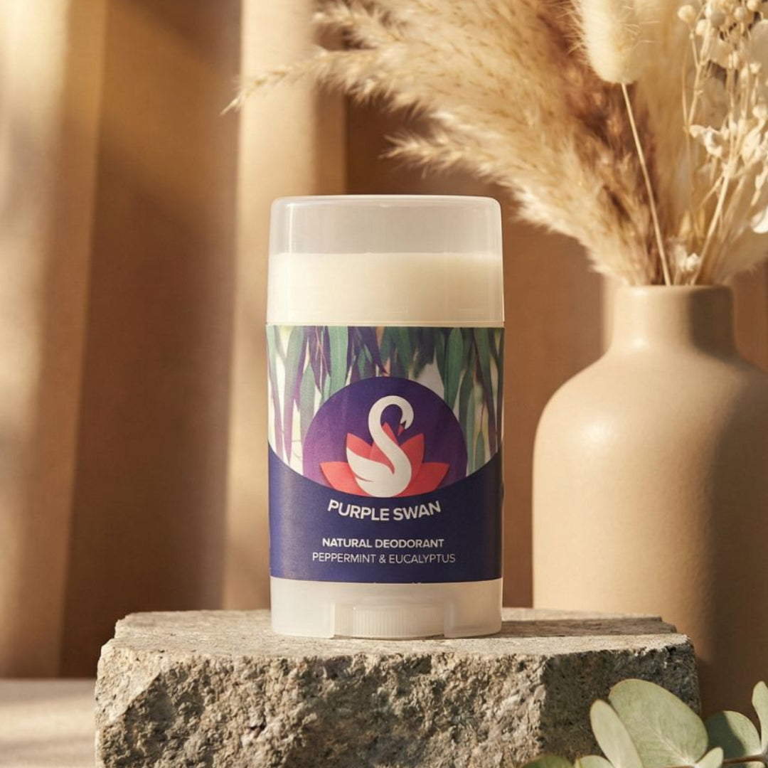 Purple Swan natural deodorant stick on a stone block with decorative elements in the background